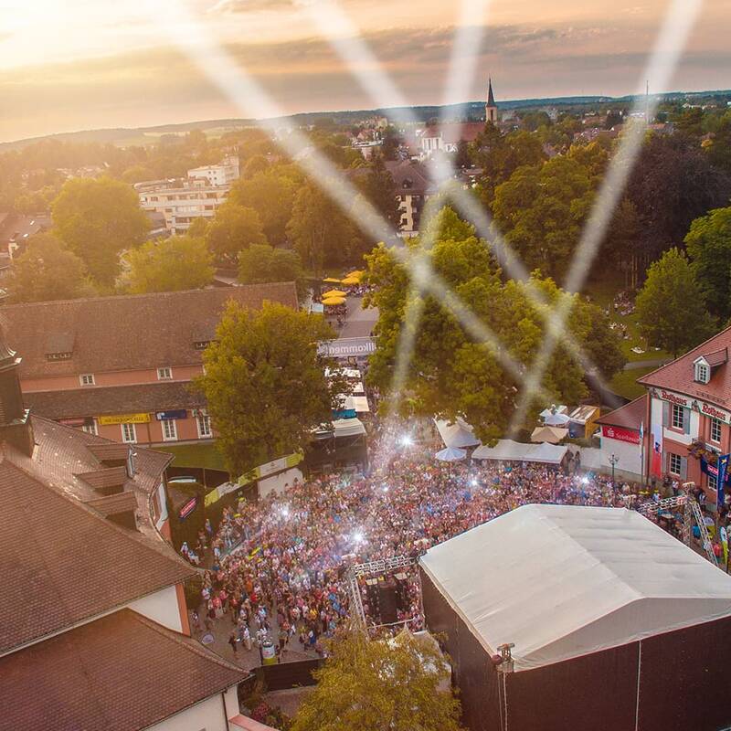 Luftaufnahme eines Festivals mit Bühnenzelt, vielen Besuchern und hellen Scheinwerfern über dem Gelände