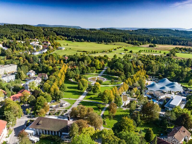 Luftaufnahme eines Parks mit Wegen, Bäumen und Gebäuden, umgeben von Feldern und Wald im Hintergrund.