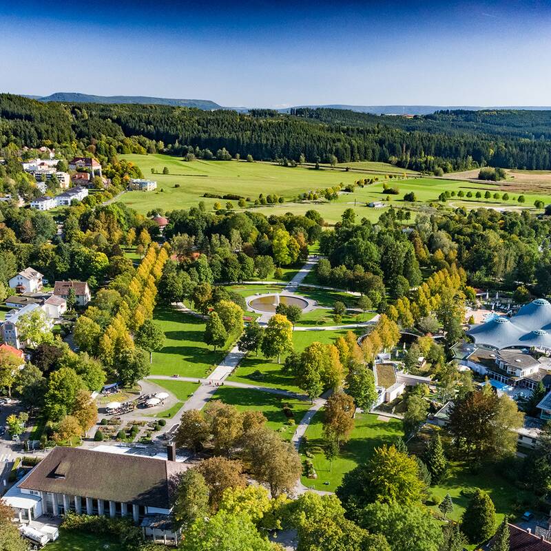 Luftaufnahme eines Parks mit Wegen, Bäumen und Gebäuden, umgeben von Feldern und Wald im Hintergrund.