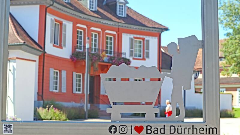 Metallschild in einem Metallrahmen und dahinter ein Gebäude mit bepflanztem Balkon