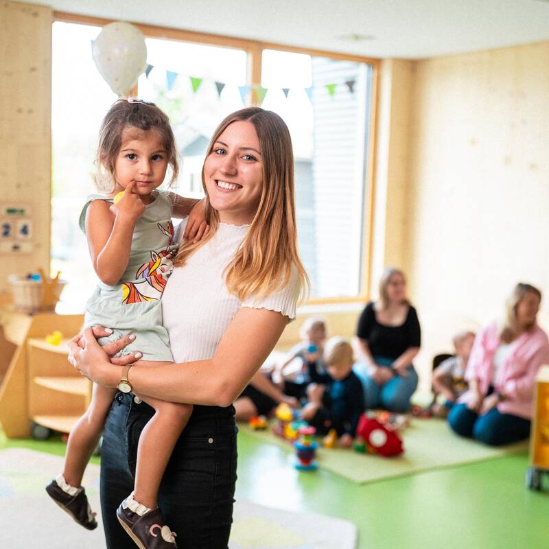 Ein Junge Frau steht im Grupenraum einer Kinderbetreuungseinrichtung und hat ein Kind auf dem Arm. Beide schauen in die Kamera. Im Hintergrund sind man mehrere erwachsene Personen und Kinder auf dem Boden zwischen Spielzeugen sitzen.