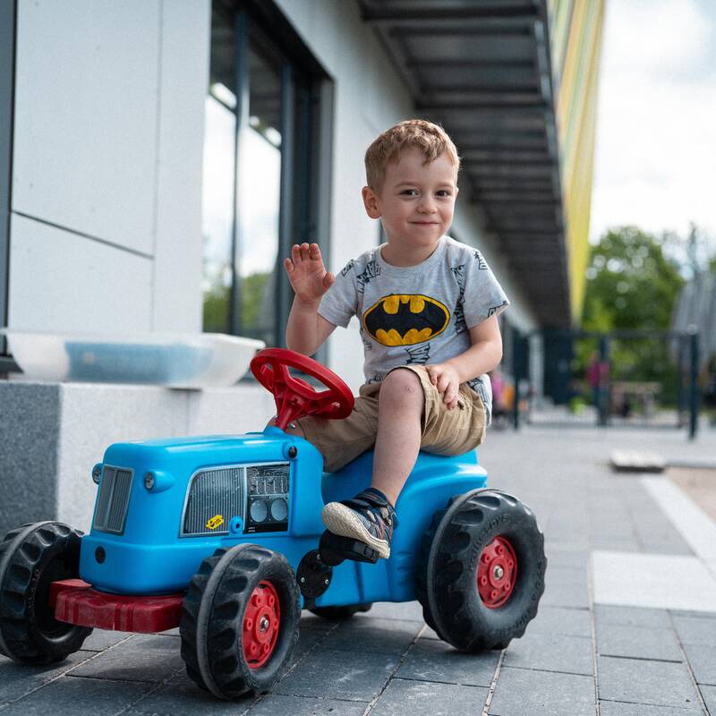Ein kleiner Junge sitzt auf einem Spielzeugtraktor im Außenbereich einer Kinderbetreuungseinrichtung und schaut winkend in die Kamera.