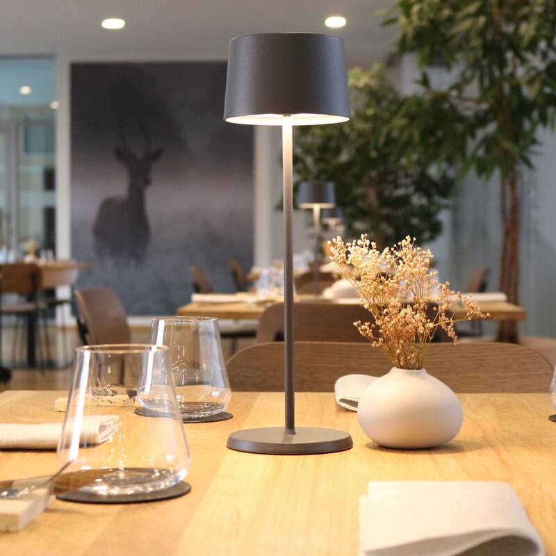 Tisch mit grauer Lampe, weißer Vase mit Trockenblumen und Gläsern in einem modernen Restaurant.