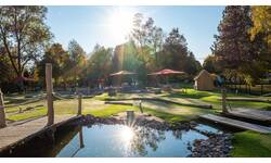 Adventure-Golf-Anlage mit mehreren Bahnen, kleinen Brücken und Sonnenschirmen, umgeben von Bäumen im Sonnenlicht.