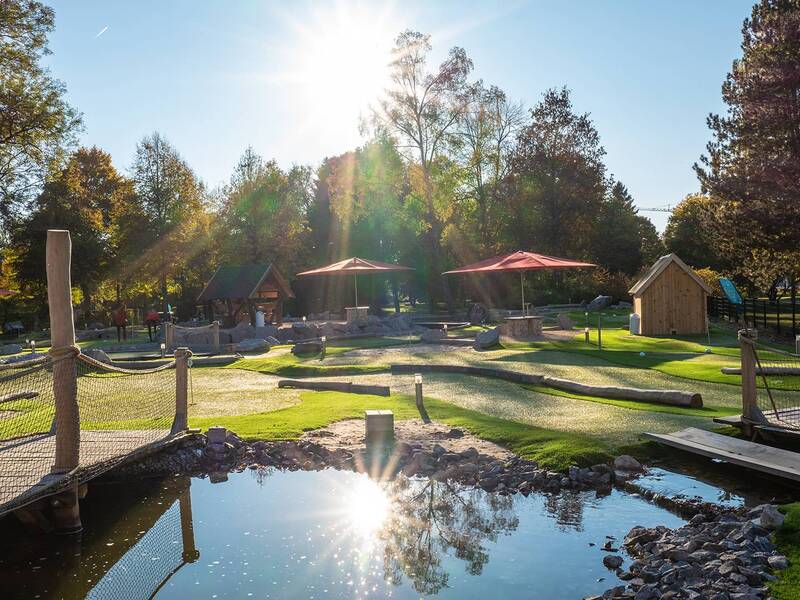 Adventure-Golf-Anlage mit mehreren Bahnen, kleinen Brücken und Sonnenschirmen, umgeben von Bäumen im Sonnenlicht.