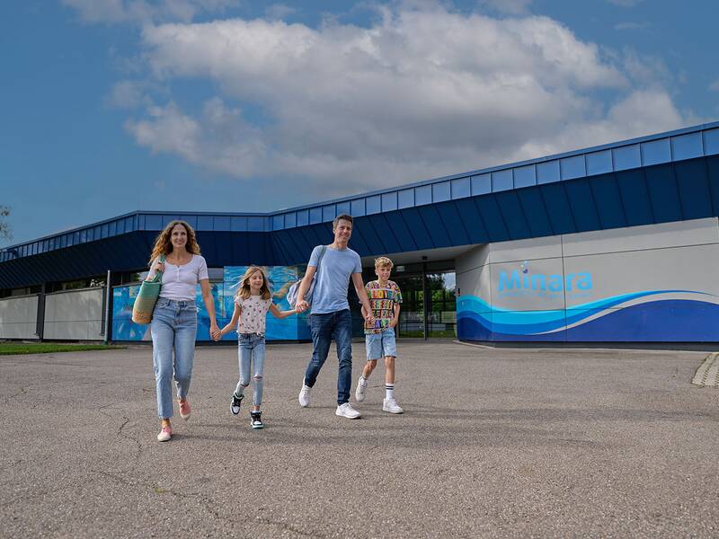 Familie mit zwei Kindern geht vor dem Freizeitbad Minara, das blaue Wellenmuster an der Wand zeigt.