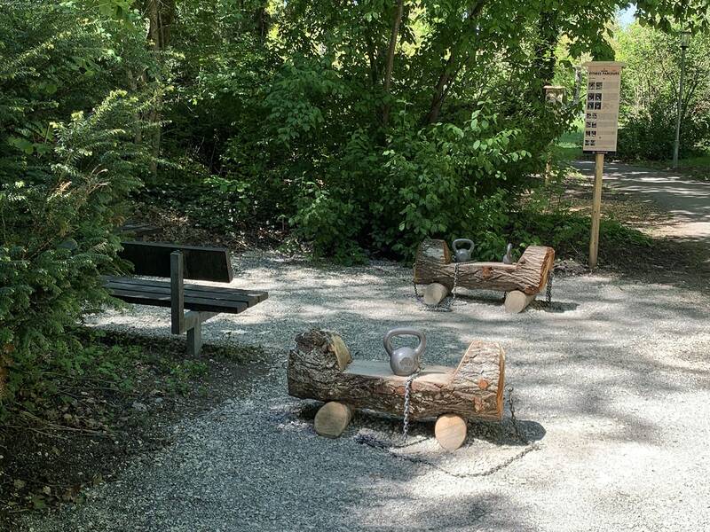 Zwei Holzbänke mit eingelassenen Kettlebells auf Kiesweg, umgeben von Büschen und Bäumen, daneben eine Parkbank und ein Schild.