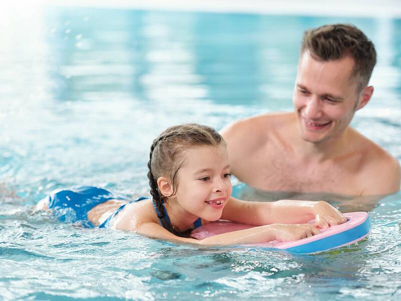 Kind mit Schwimmhilfe im Wasser, begleitet von einem Erwachsenen, der hinter dem Kind schwimmt.