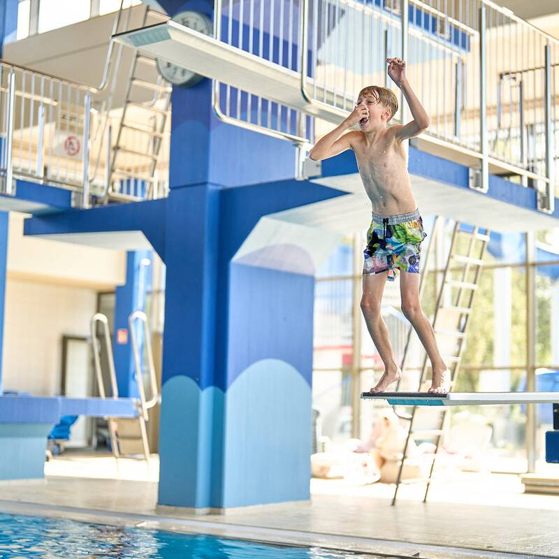 Junge springt vom Sprungbrett in ein Hallenbad mit blauen Säulen und großen Fenstern