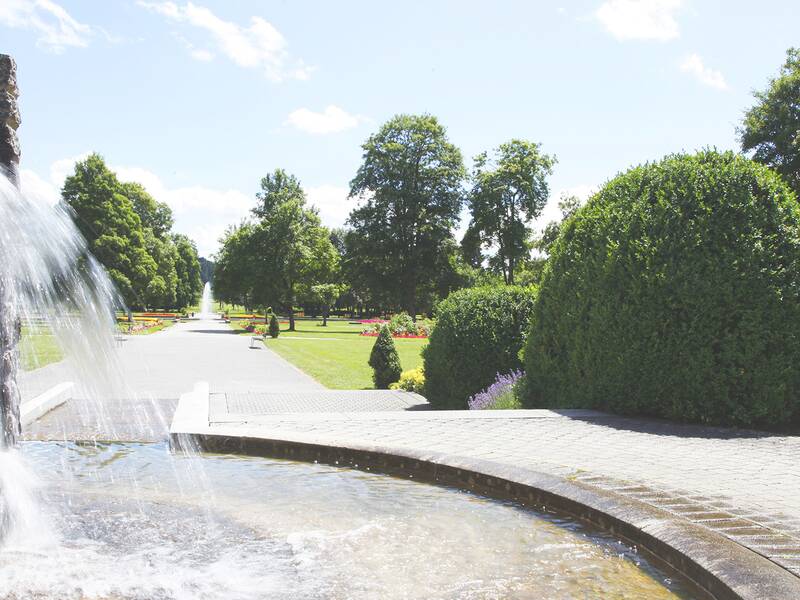 Springbrunnen mit Wasserfall im Kurpark mit gepflastertem Weg, Büschen und Bäumen unter blauem Himmel.