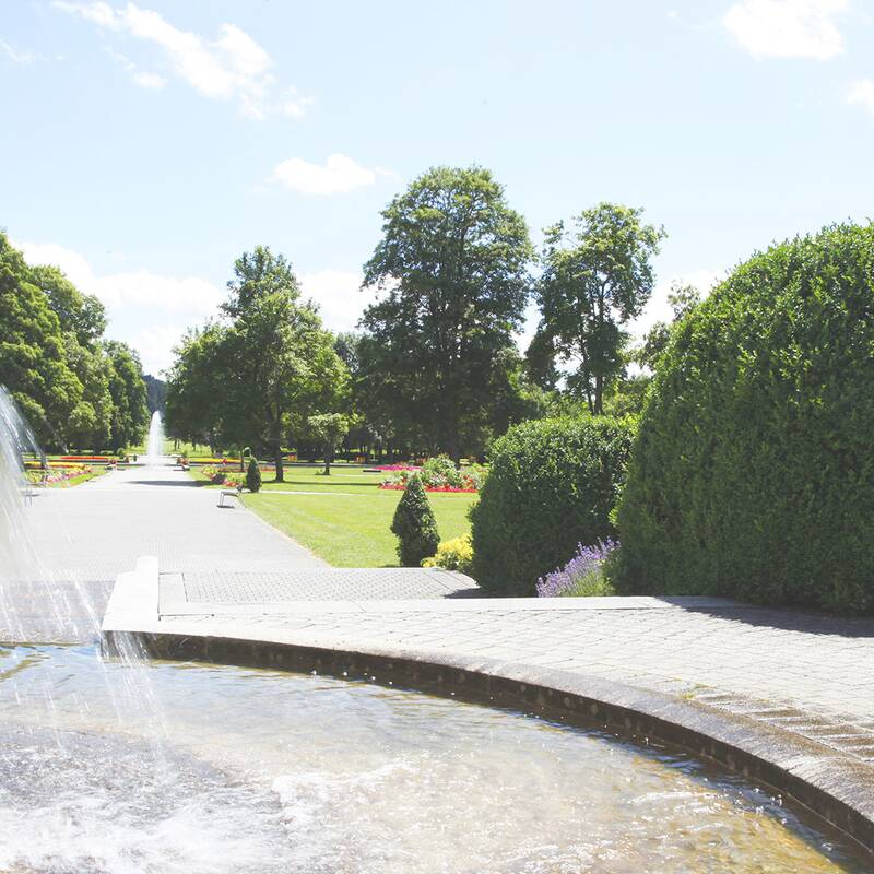 Springbrunnen mit Wasserfall im Kurpark mit gepflastertem Weg, Büschen und Bäumen unter blauem Himmel.