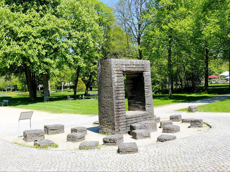 Steinrahmen mit mehreren kleinen Steinblöcken auf gepflastertem Platz in einem Park mit Bäumen und Wegen