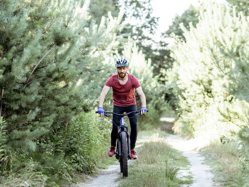 Person in rotem T-Shirt und schwarzer Hose fährt Fahrrad auf einem Waldweg, umgeben von grünen Bäumen und Sträuchern.