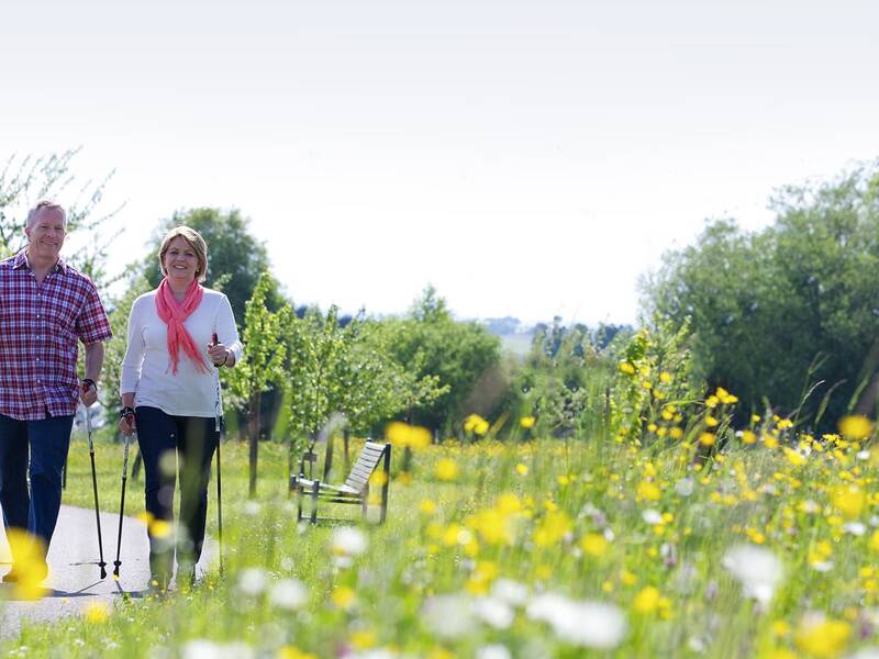 Zwei Personen gehen mit Nordic-Walking-Stöcken auf einem Weg, umgeben von grünen Bäumen und gelben Blumen.