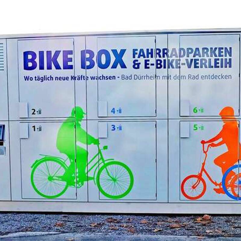 Fahrradbox mit blauer Aufschrift 'BIKE BOX FAHRRADPARKEN & E-BIKE-VERLEIH' und farbigen Silhouetten von zwei Radfahrern, einer grün, einer orange.