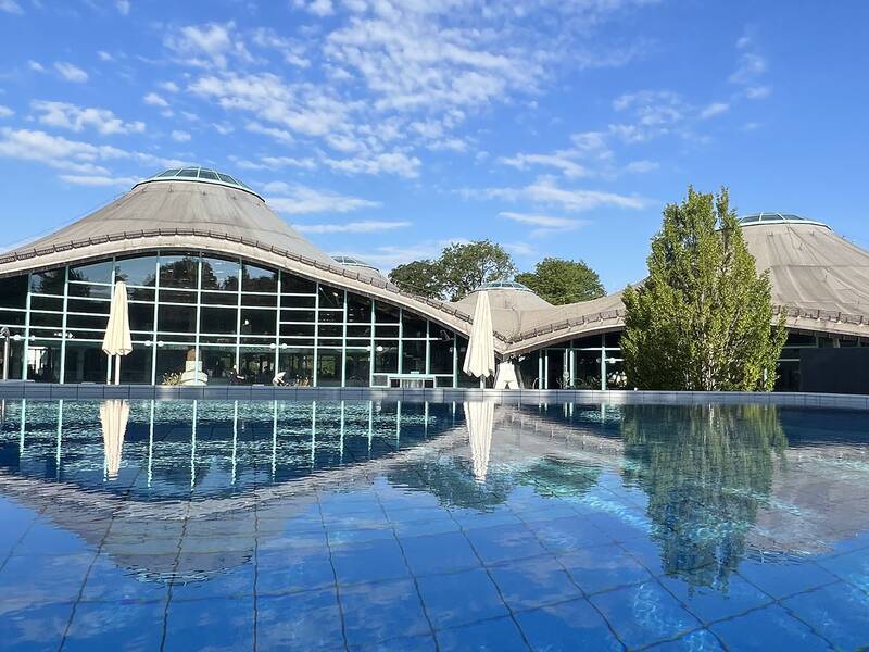 Gebäude mit gewölbtem Dach und großer Glasfront, davor ein Schwimmbecken mit Wasserspiegelung, blauer Himmel.