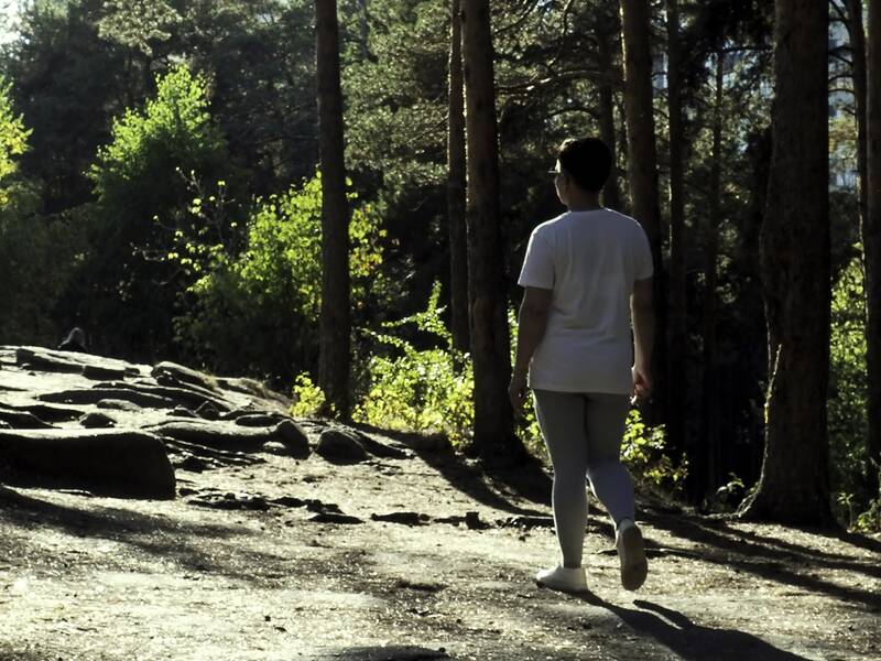 Frau in weißem T-Shirt und grauer Leggings geht auf einem Waldweg zwischen hohen Kiefern.