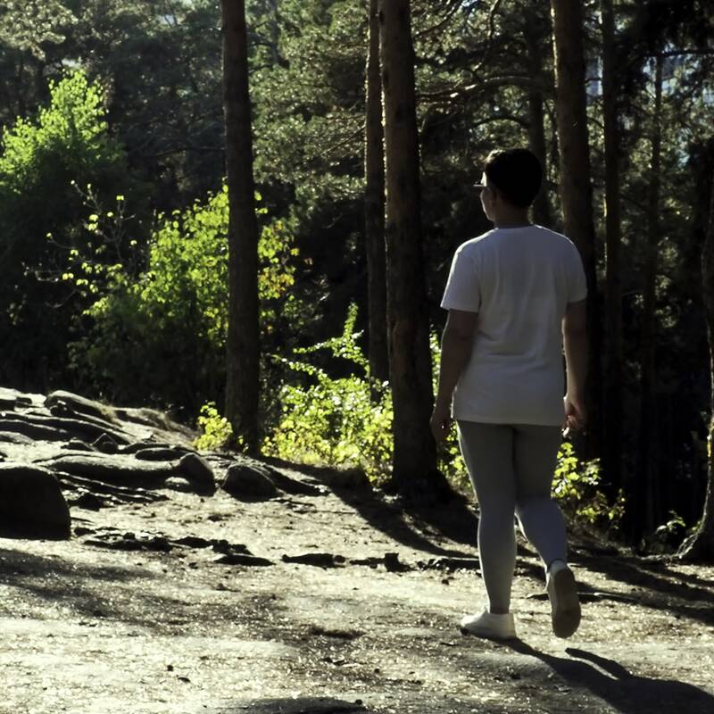Frau in weißem T-Shirt und grauer Leggings geht auf einem Waldweg zwischen hohen Kiefern.