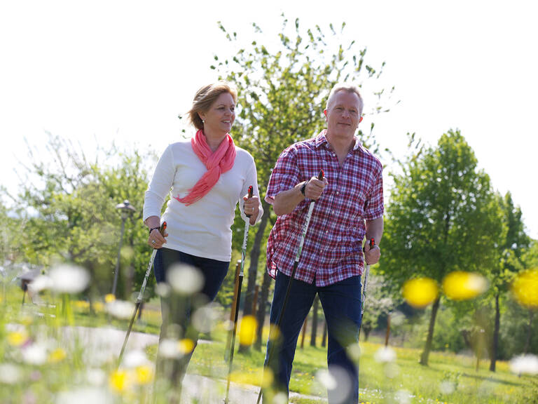 Zwei Personen beim Nordic Walking in einem Park, eine trägt ein weißes Oberteil mit pinkem Schal, die andere ein rot-kariertes Hemd.