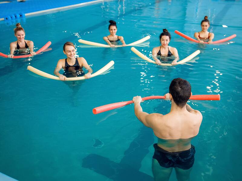 Mann im Schwimmbad hält rote Poolnudel, vor ihm fünf Frauen im Wasser mit weißen und roten Poolnudeln, alle in Badebekleidung.