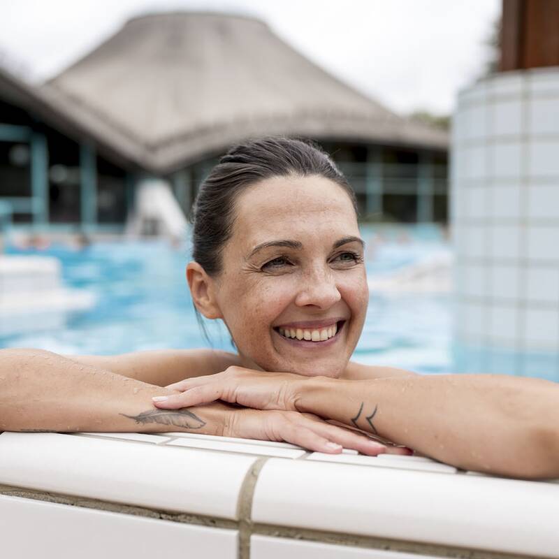 Person lehnt mit verschränkten Armen am Beckenrand eines Schwimmbeckens mit weißen Fliesen und blauem Wasser.