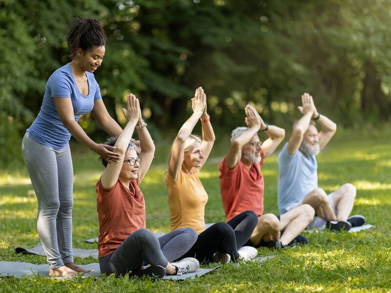 Fünf Personen sitzen auf Yogamatten auf einer Wiese, vier davon heben die Hände über den Kopf, während eine stehende Person eine davon unterstützt.