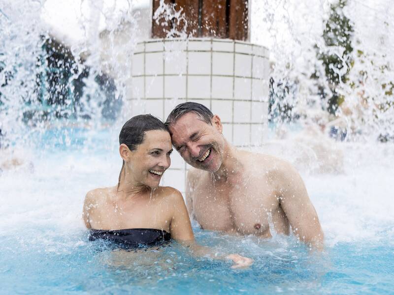 Paar im Außenbecken einer Therme unter einem Wasserfall, beide im Wasser bis zur Brust sichtbar.