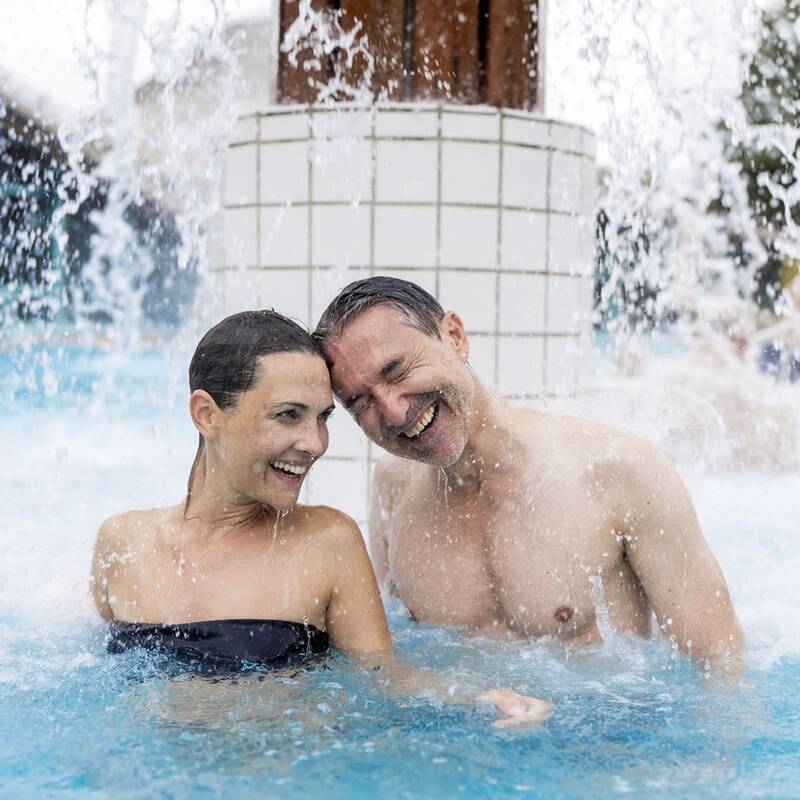 Paar im Außenbecken einer Therme unter einem Wasserfall, beide im Wasser bis zur Brust sichtbar.