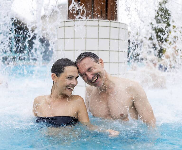 Paar im Außenbecken einer Therme unter einem Wasserfall, beide im Wasser bis zur Brust sichtbar.