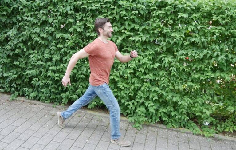 Junger Mann in orangefarbenem T-Shirt und blauer Jeans geht auf einem gepflasterten Weg vor einer grünen Hecke.