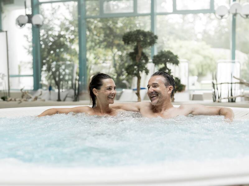 Zwei Personen lehnen entspannt in einem Whirlpool in einem hellen, begrünten Innenraum.