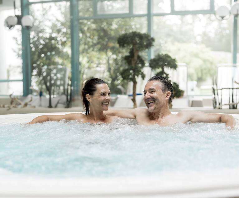 Zwei Personen lehnen entspannt in einem Whirlpool in einem hellen, begrünten Innenraum.