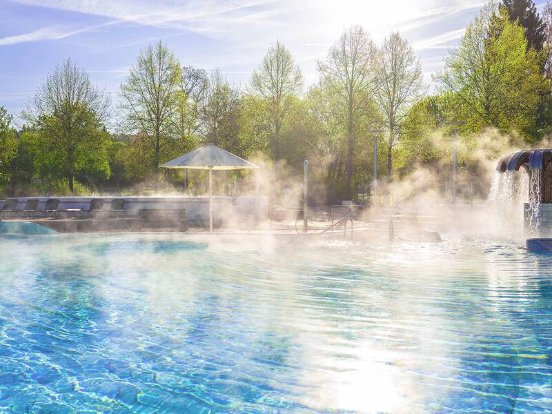 Dampfendes Außenbecken mit Wasserpilz und Liegestühlen im Hintergrund, umgeben von Bäumen.
