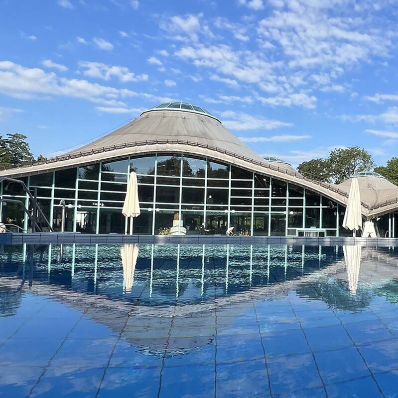 Gebäude mit gewölbtem Dach und großer Glasfassade spiegelt sich in einem ruhigen Wasserbecken
