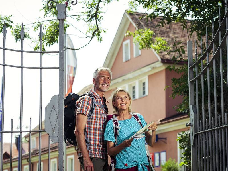 Zwei Personen mit Rucksäcken stehen vor einem offenen Tor, eine hält einen Stadtplan, im Hintergrund das Haus des Gastes..