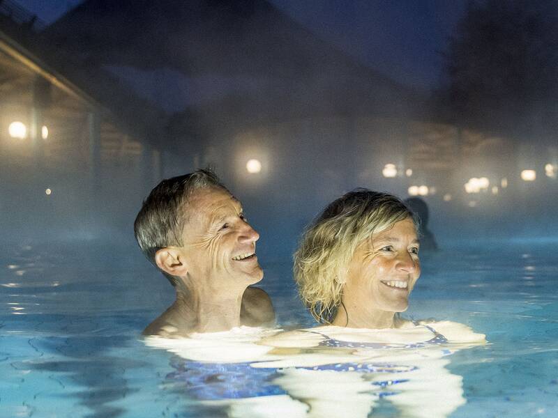 Zwei Personen im Außenpool bei Nacht, umgeben von Dampf und beleuchteten Solemar im Hintergrund.