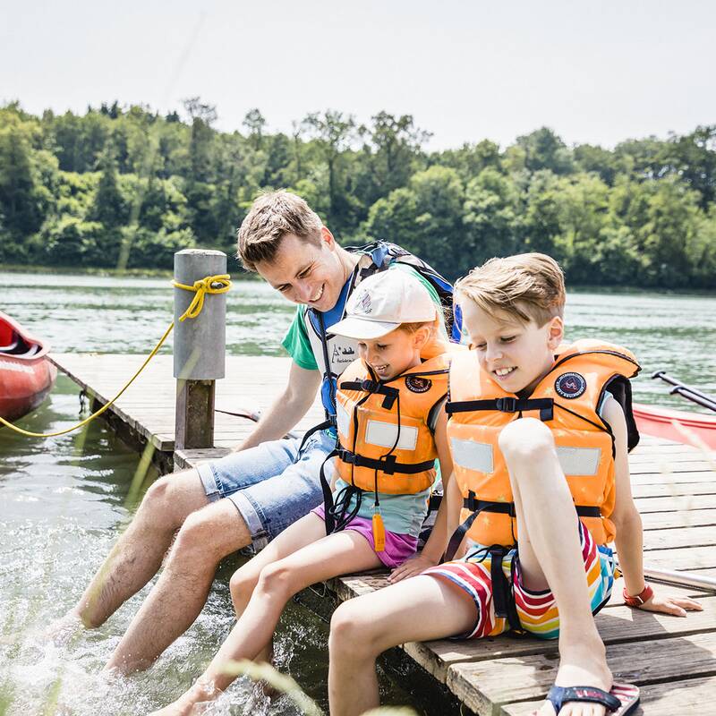Zwei Kinder und ein Erwachsener mit Schwimmwesten sitzen am Steg, neben einem roten Kanu auf dem Wasser.