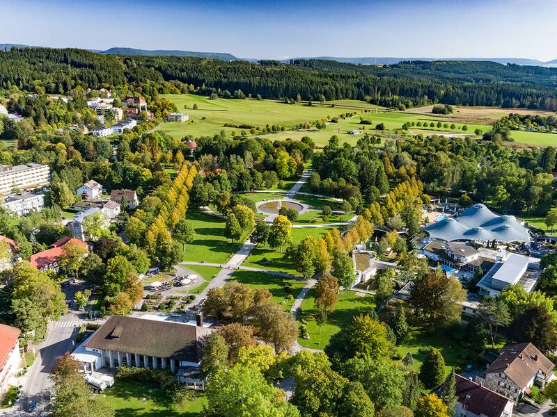 Luftaufnahme des Kurparks mit vielen Bäumen, Wegen und Gebäuden, umgeben von Feldern und Wald im Hintergrund.