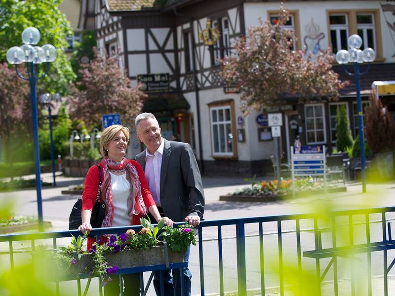Ein Paar steht an einem blauen Geländer mit Blumenkasten in einer Straße mit Fachwerkhäusern und Straßenlaternen im Hintergrund.