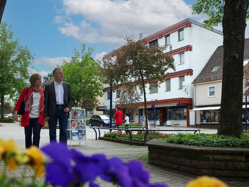 Zwei Personen gehen auf einem gepflasterten Weg in einer Innenstadt mit Bäumen, Blumen und mehrstöckigen Gebäuden im Hintergrund.