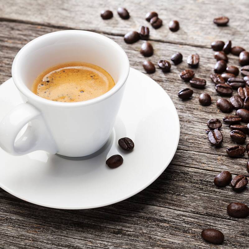 Weiße Espressotasse mit Schaum auf Untertasse, umgeben von verstreuten Kaffeebohnen auf Holzoberfläche.