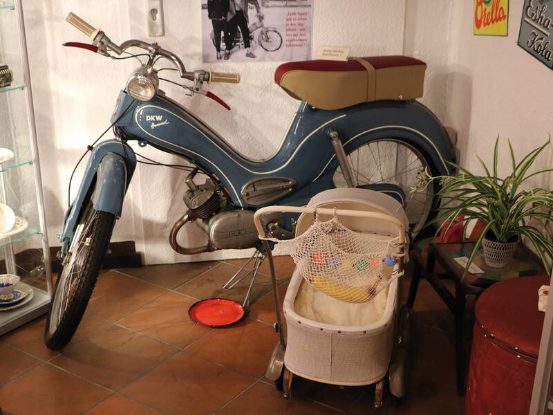 Altes blaues Motorrad mit braunem Sitz steht in einem Museum neben einem weißen Kinderwagen und Pflanzen.