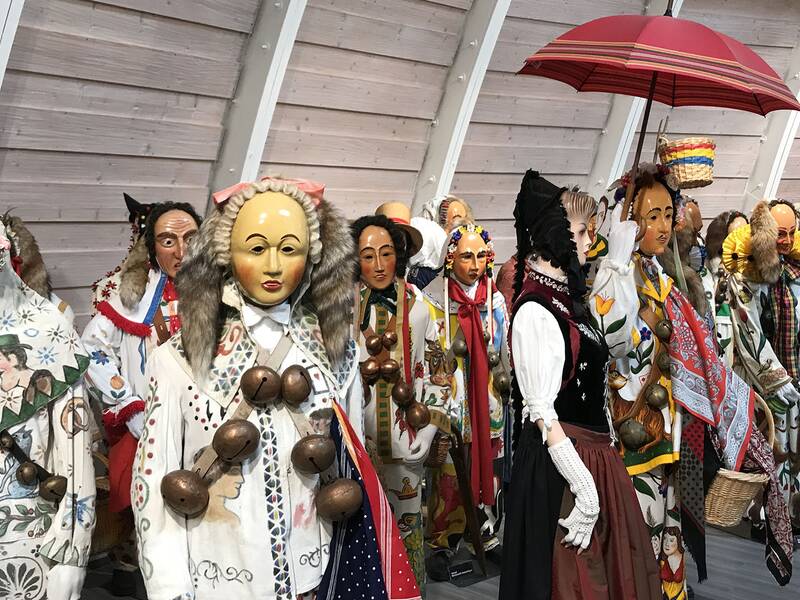 Figuren in traditionellen, farbenfrohen Kostümen mit großen Glocken und einem roten Regenschirm in einer Halle mit Holzverkleidung.