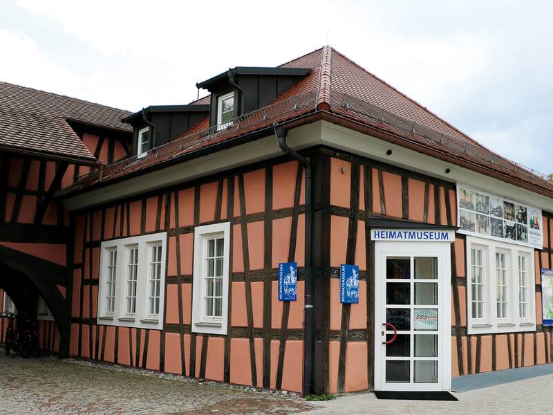 Fachwerkhaus mit Schildern, darunter ein Schild mit der Aufschrift 'Heimatmuseum', an einer gepflasterten Straße mit parkenden Autos.
