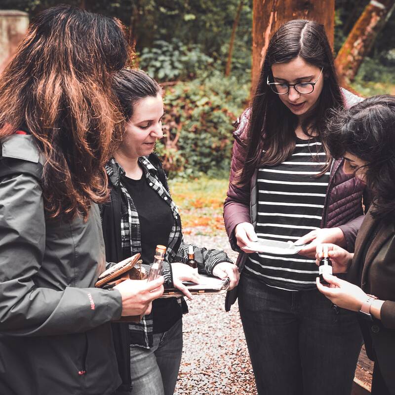 Vier Frauen stehen im Freien auf einem Kiesweg, halten kleine Gegenstände und lösen ein Rätsel.