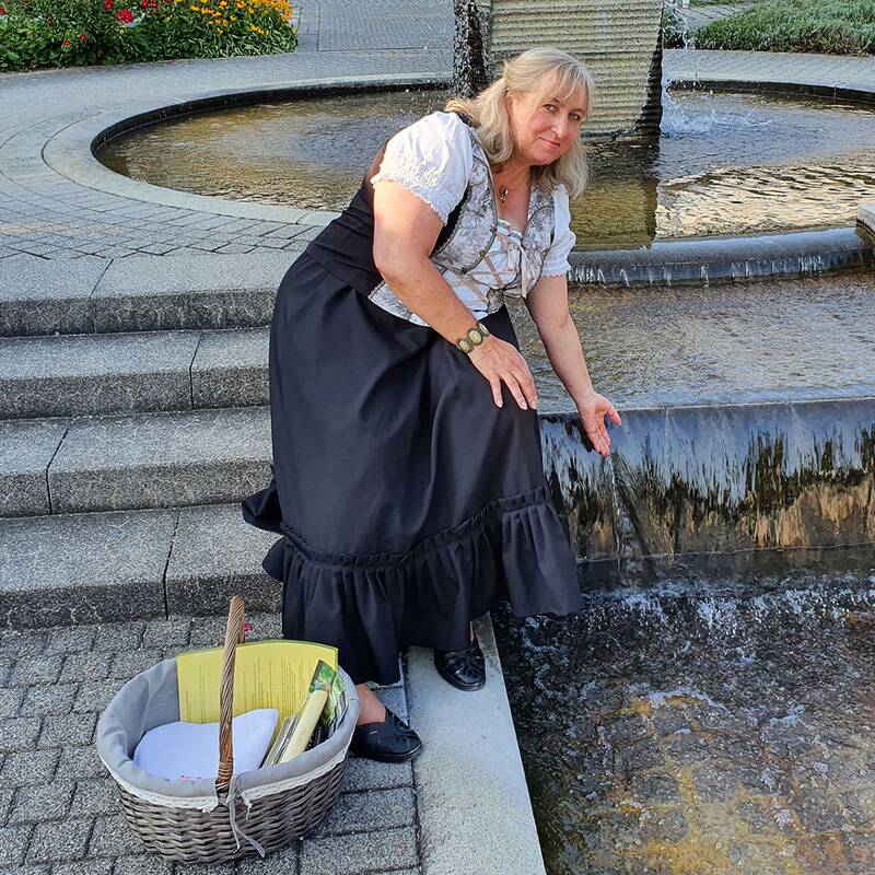 Frau in traditioneller Tracht kniet an einem Wasserbecken mit fließendem Wasser, daneben ein Korb.