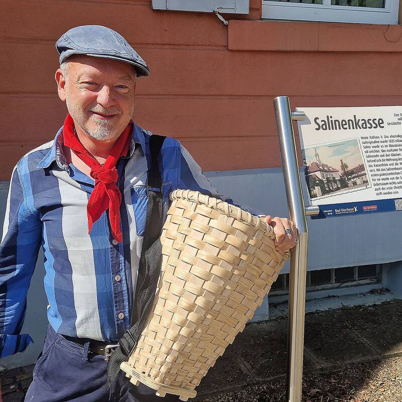 Mann mit Schiebermütze, rotem Halstuch und blauem Hemd hält einen großen geflochtenen Korb vor einem Gebäude mit Infotafel zur Saline.