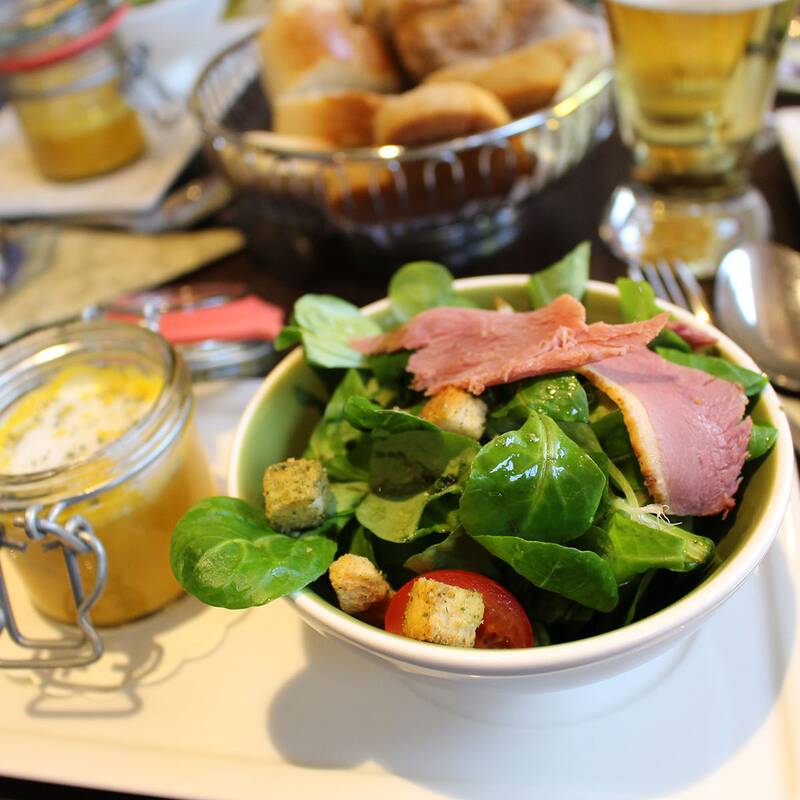 Schüssel mit grünem Salat, Tomaten, Croutons und Scheiben von rosa Fleisch auf einem weißen Tablett mit kleinem Glas mit Sauce und Brotkorb im Hintergrund.