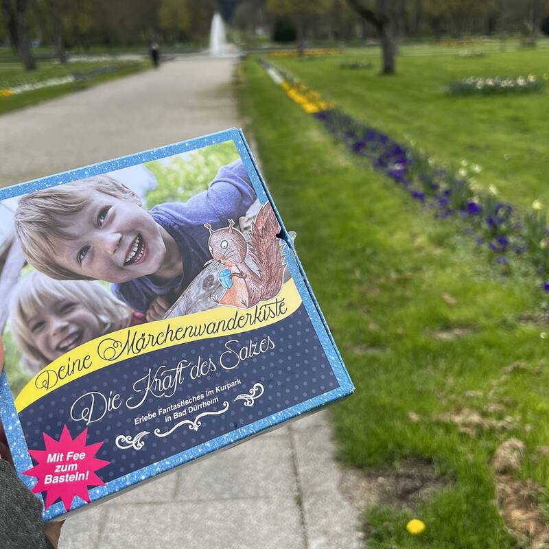 Hand hält eine bunte Schachtel mit dem Titel 'Deine Märchenwanderkiste Die Kraft des Salzes' vor einem Parkweg mit Blumenbeeten und Bäumen.