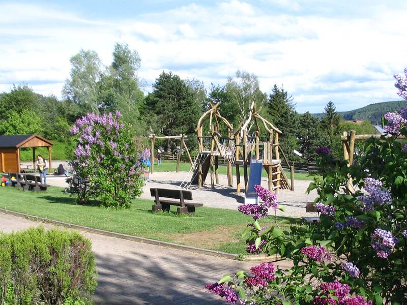 Spielplatz mit Holzspielgeräten, Bänken und blühenden Fliedersträuchern in einer grünen Parklandschaft unter blauem Himmel.